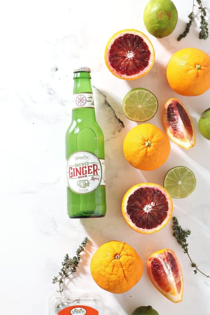 A ginger beer is surrounded by blood oranges, limes and fresh thyme on a marble background