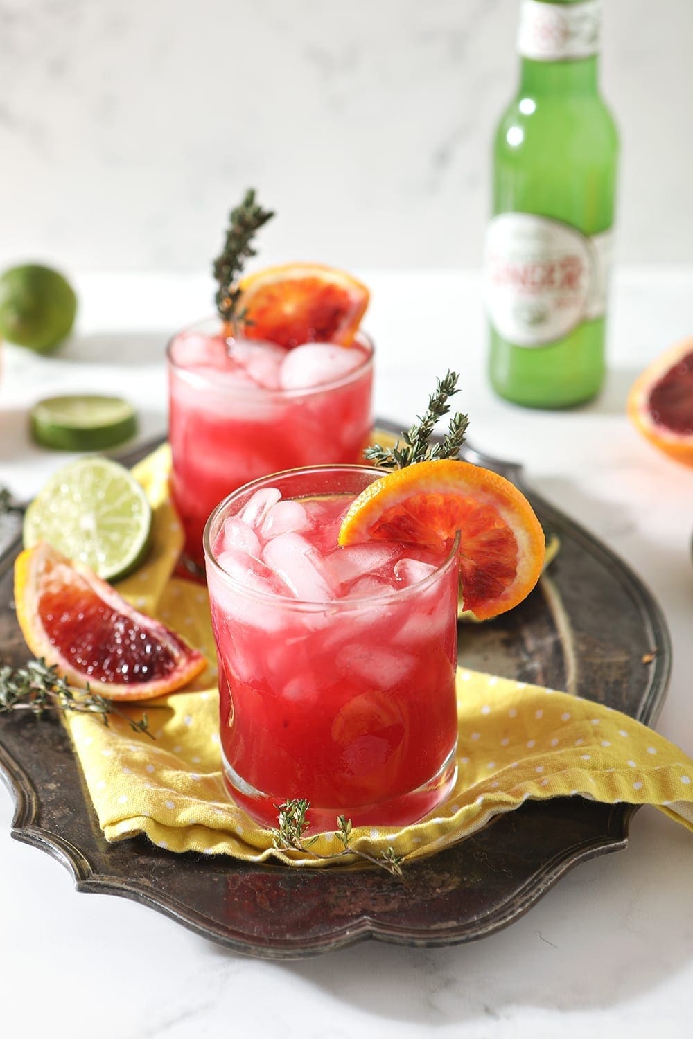 Two Blood Orange Mocktail Mules sit on a yellow napkin on a silver platter, surrounded by sliced citrus