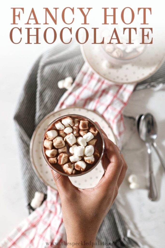 A woman holds a mug of gourmet hot chocolate, from above, with marshmallows on top and Pinterest text