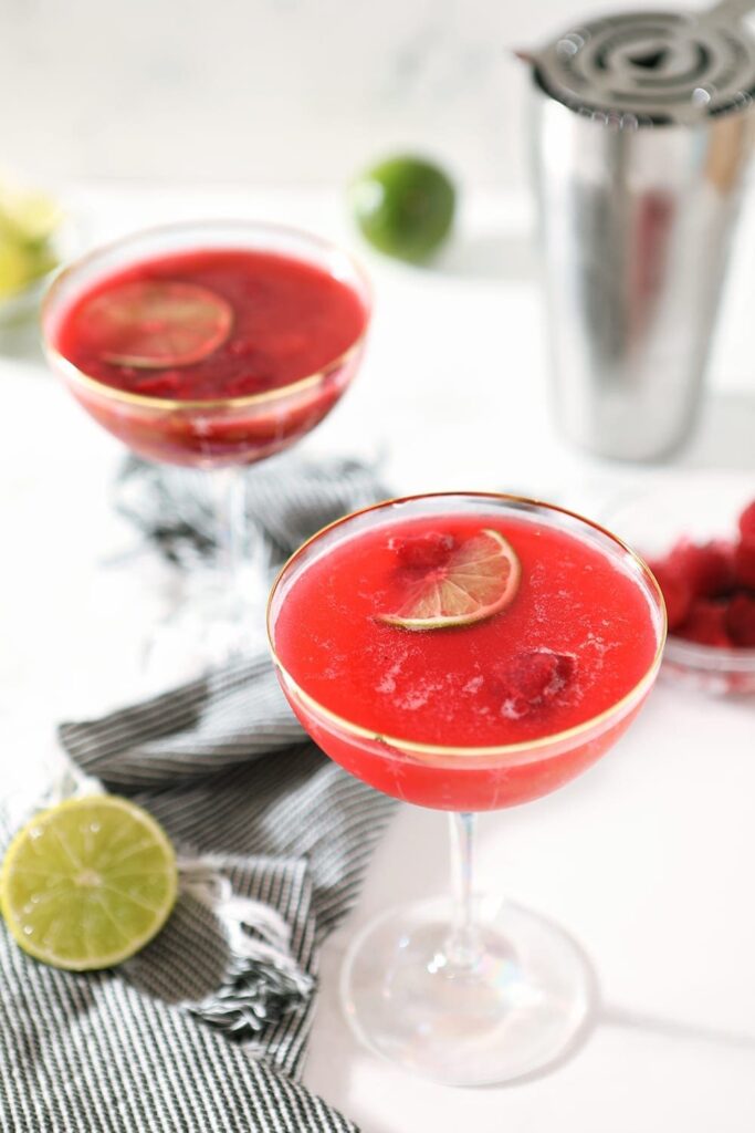 Two glasses of Raspberry Moscow Mules sit on a marble countertop, with a shaker and limes