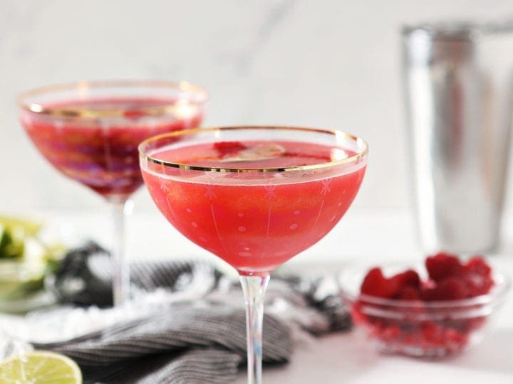 Two Raspberry Mules are shown on a marble background, with a striped blue cloth, limes and raspberries