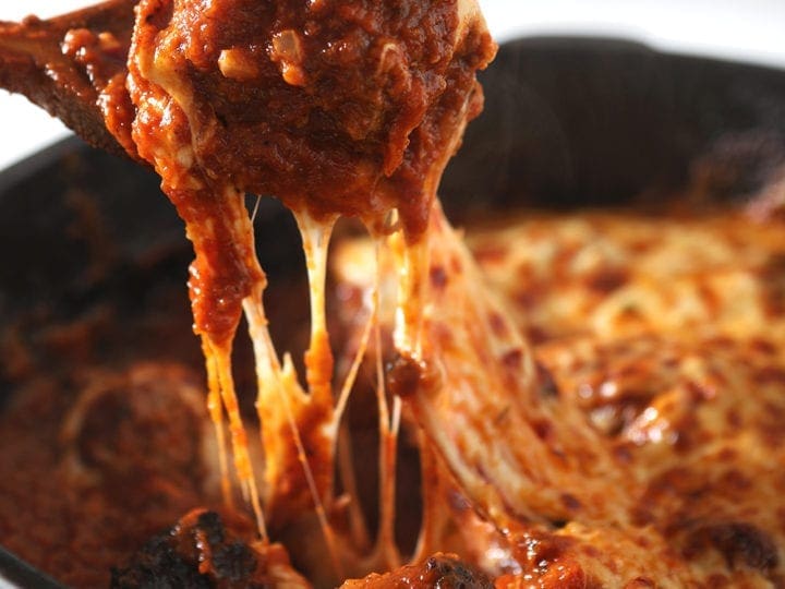 A wooden spoon pulls a meatball out of the skillet, with a cheese pull