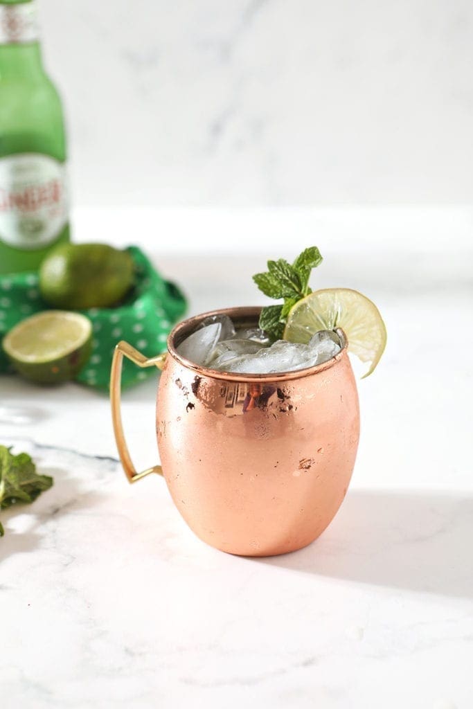 A copper mug holding an Irish Whiskey Moscow Mule sits on a marble background, surrounded by ingredients