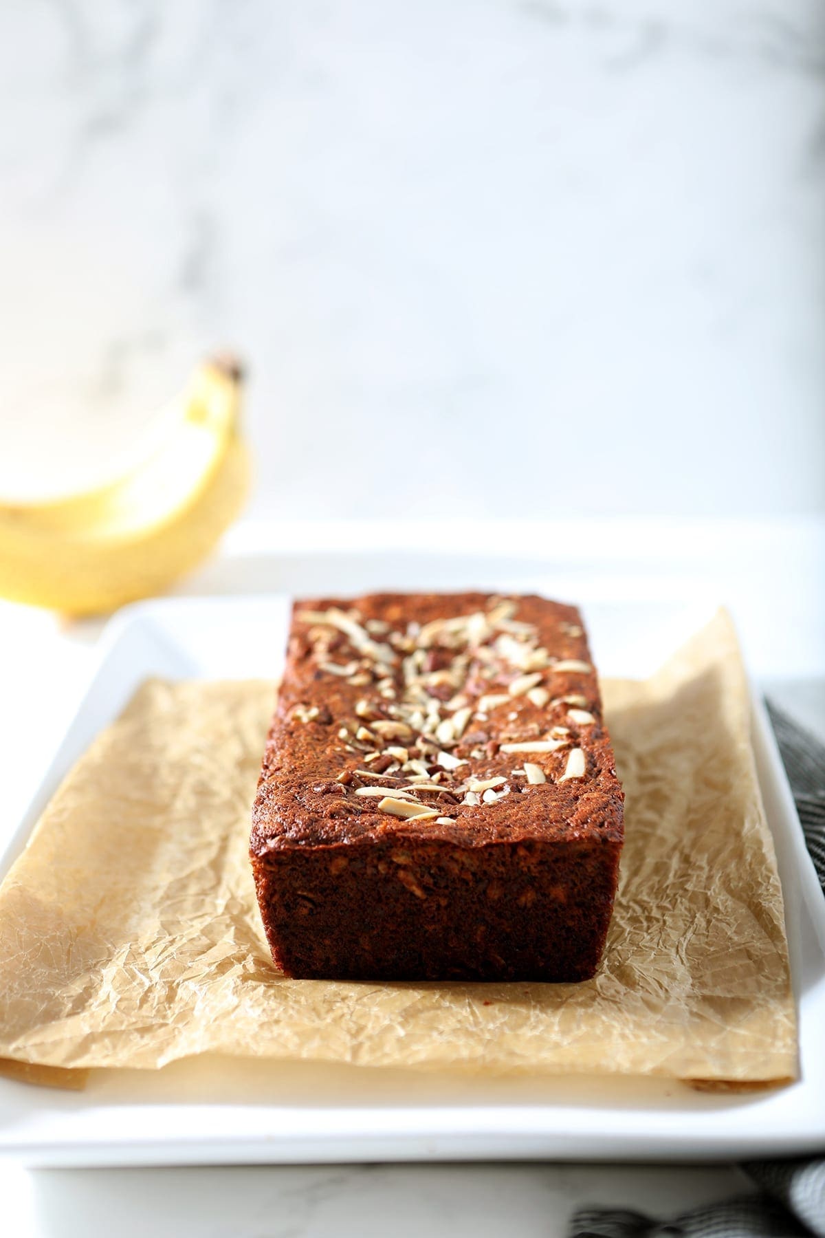 A loaf of Banana Bread sits on a piece of parchment paper on a white platter, ready for slicing