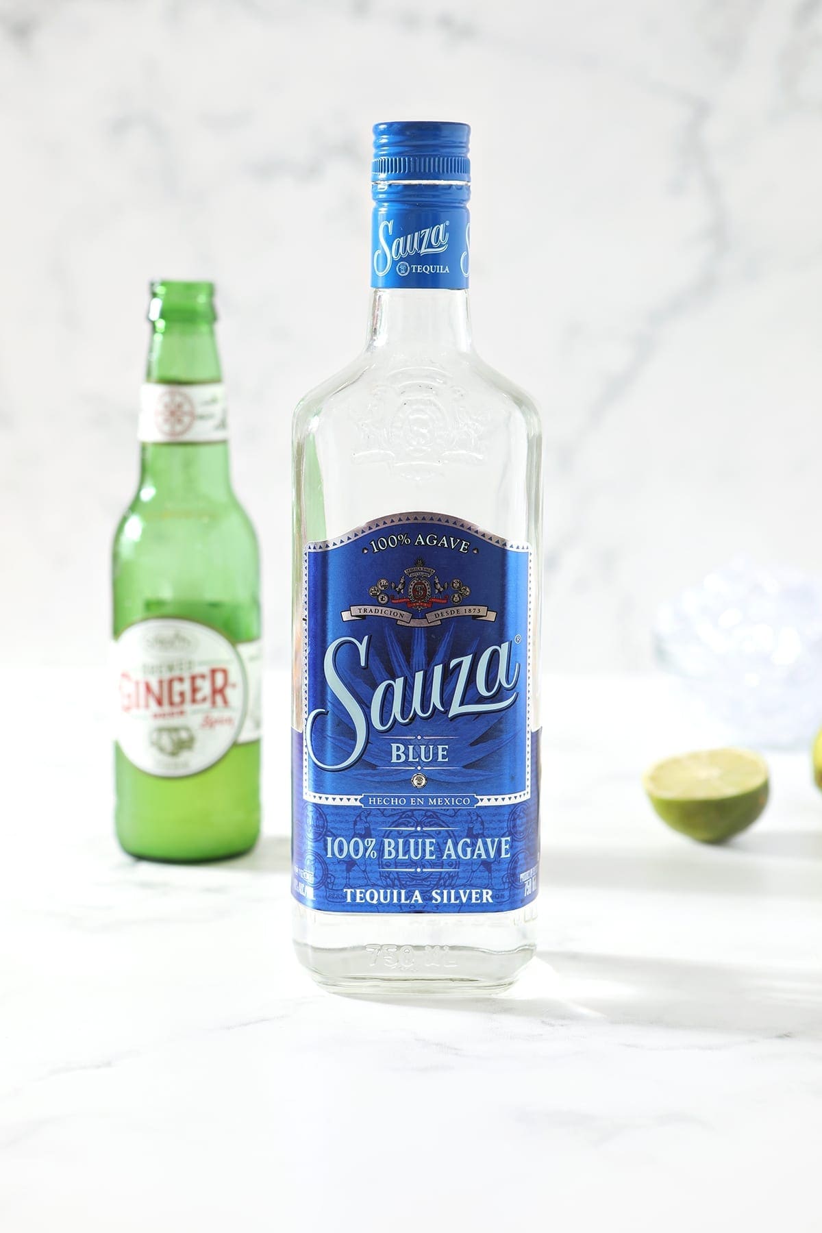 A bottle of tequila, a bottle of ginger beer and a half of a lime are shown on a marble countertop