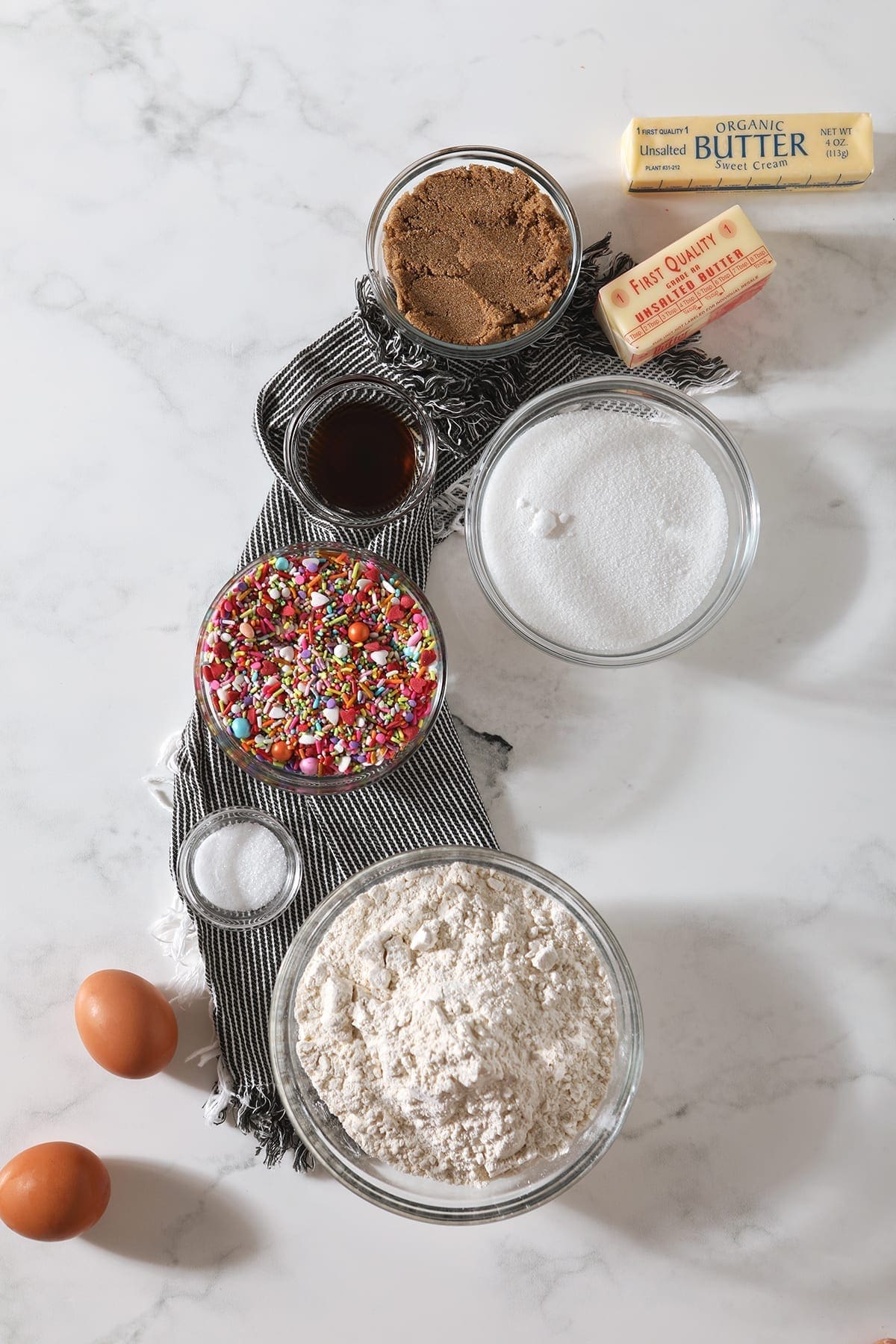 Flour, eggs, sprinkles, sugars, butter and more from above on a marble tabletop