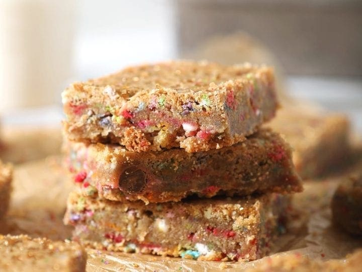 A stack of three cookie bars, shown on brown paper with other bars and in front of a glass of milk and a baking sheet