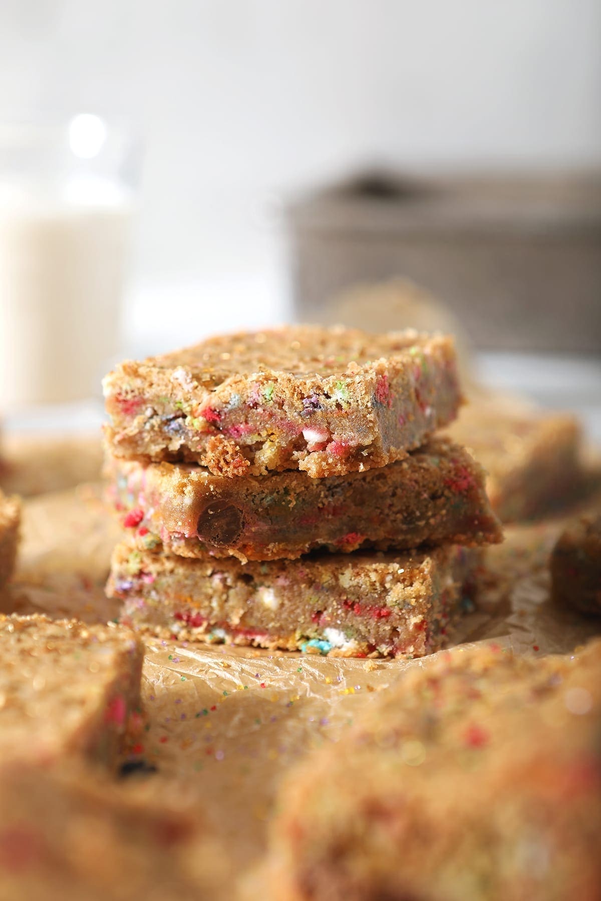 A stack of three cookie bars, shown on brown paper with other bars and in front of a glass of milk and a baking sheet