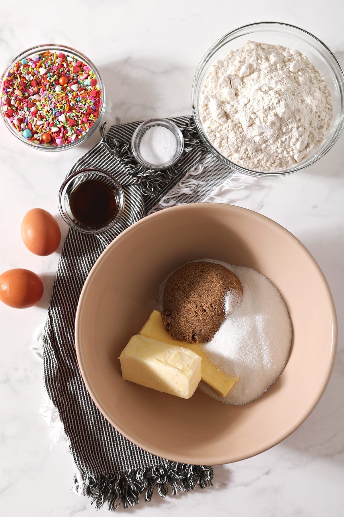 Sticks of butter and sugars are shown in a tan bowl, with eggs, vanilla and other ingredients around them