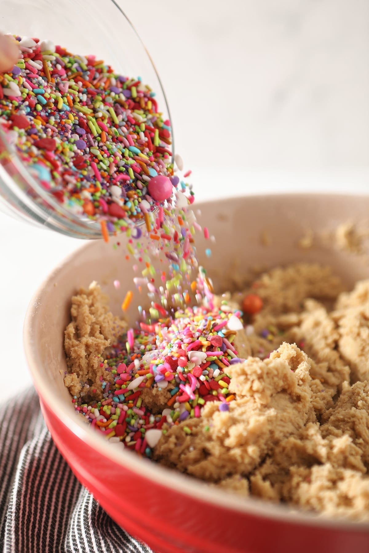 Sprinkles are poured into a red bowl of cookie dough