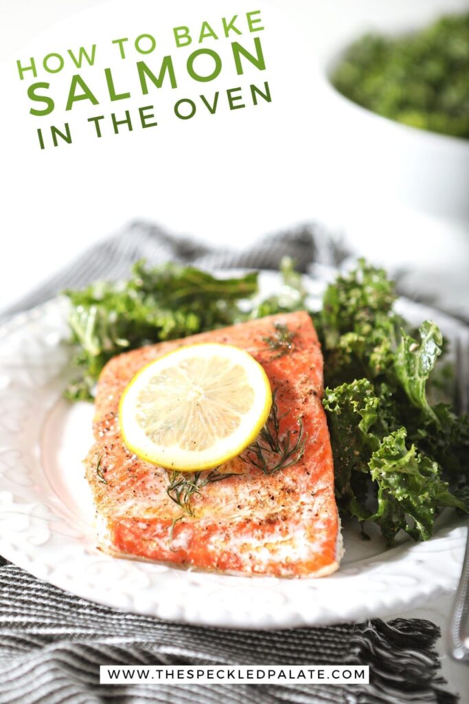 A baked salmon filet with a lemon round on parchment paper on white plate with a kale salad and Pinterest text stating, "How to Bake Salmon in the Oven"