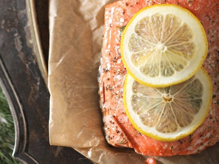 A salmon filet garnished with a lemon round is served on parchment paper over a silver platter