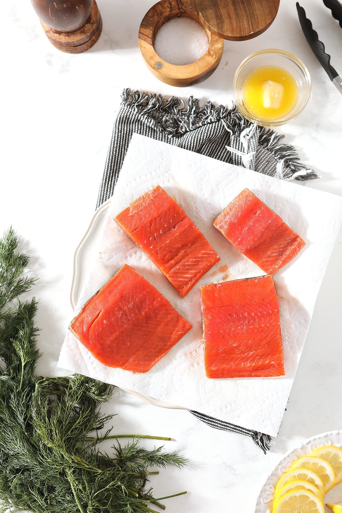 Salmon filets on a white plate lined with paper towels, surrounded by fresh dill, lemon rounds, melted butter and containers of salt and pepper