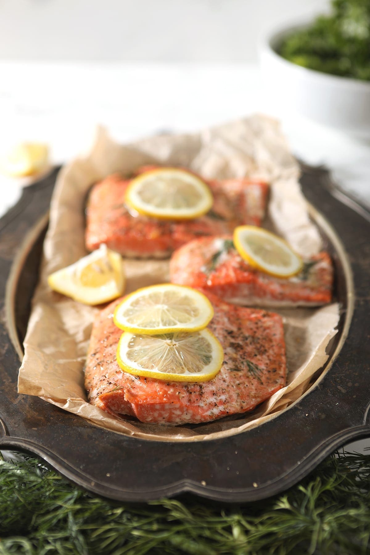 Three salmon filets on a metal platter on parchment paper, surrounded with fresh dill and garnished with lemon rounds