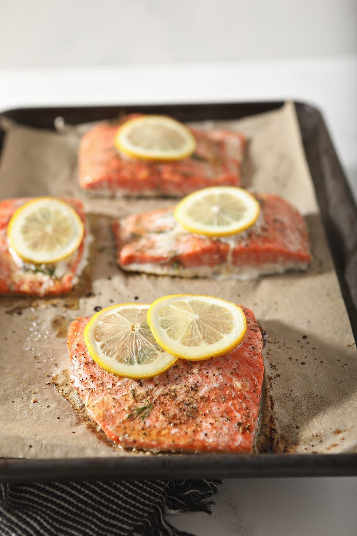 Four salmon filets sit on a baking sheet, garnished with lemon rounds, over a dark grey and white striped towel