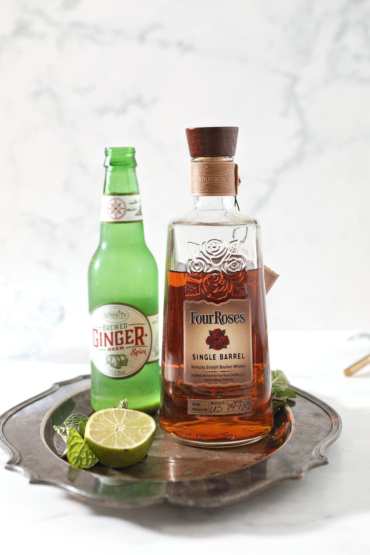 A bottle of bourbon, ginger beer and a lime with mint are shown on a silver platter