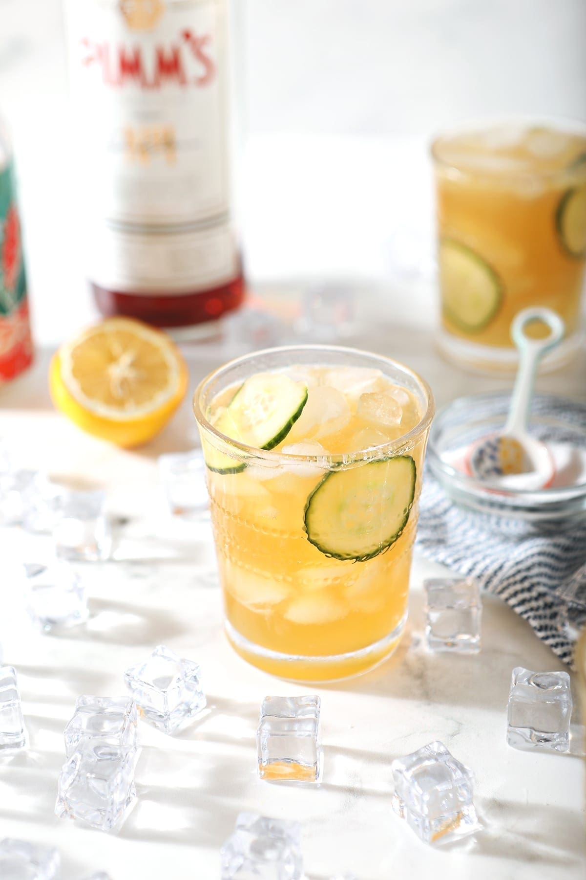 Two Pimm's Cups are surrounded by ingredients and ice, from a 45 degree angle, on marble