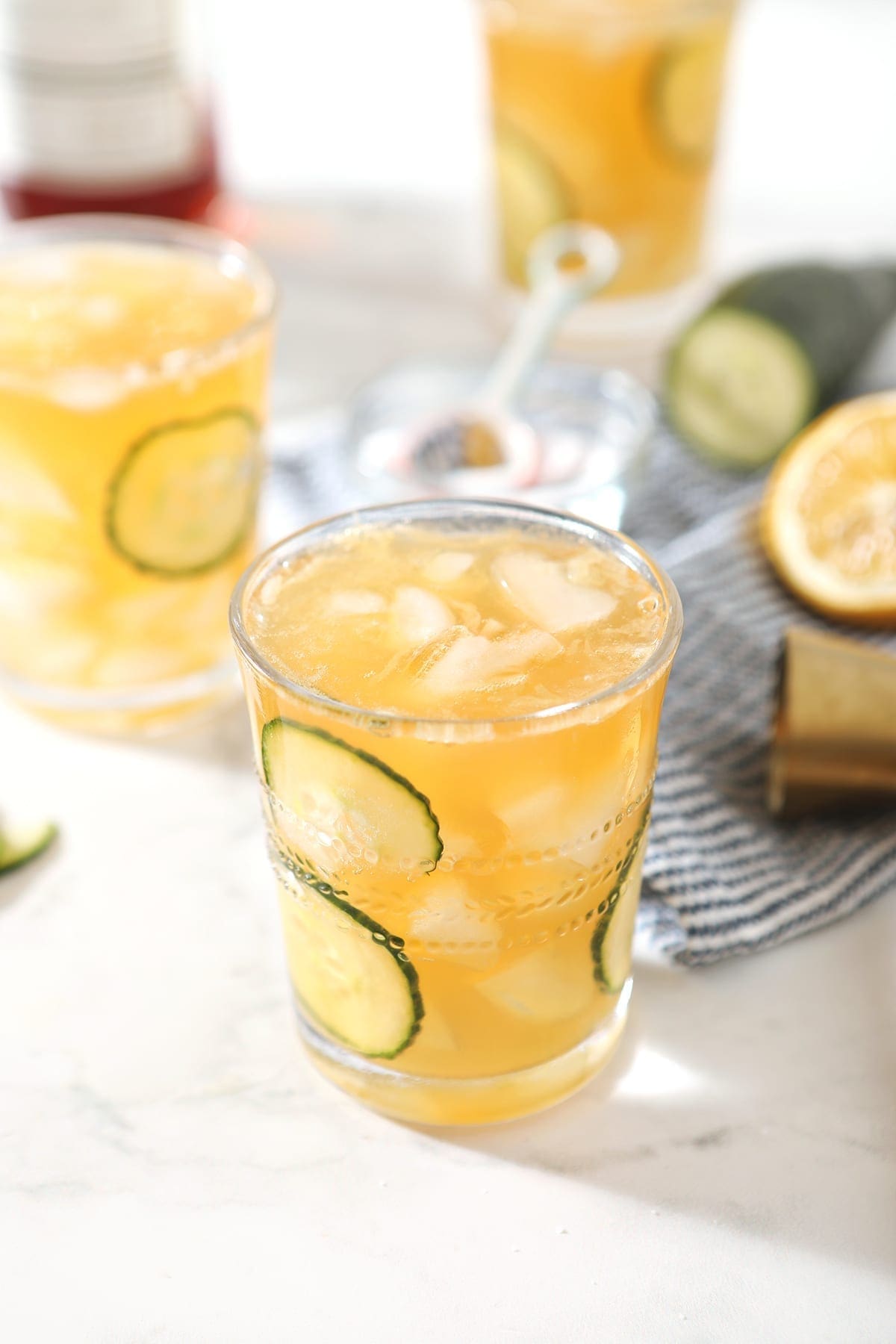 Three glasses holding Pimm's Cups are shown, surrounded by ingredients and garnished with cucumber rounds