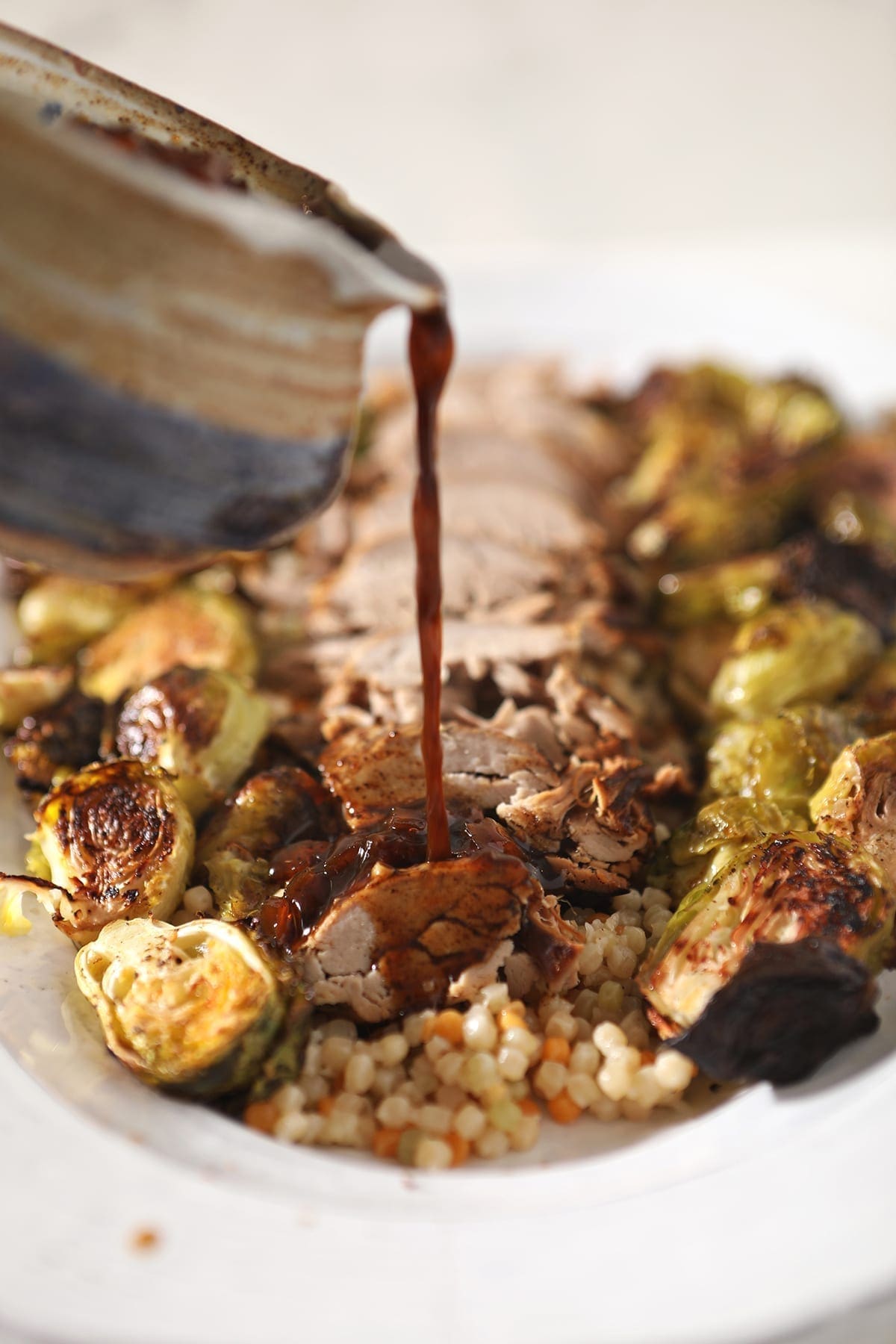 A sauce pours into a sliced pork tenderloin on a white platter with roasted brussels sprouts