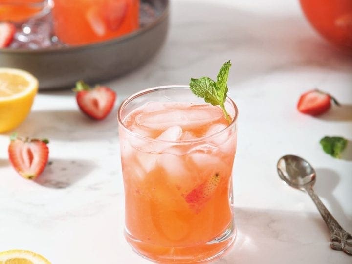 A glass of Strawberry Lemonade is shown with fresh strawberries and lemon slices