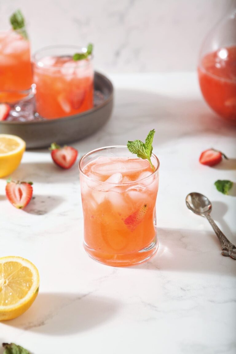 A glass of Strawberry Lemonade is shown with fresh strawberries and lemon slices