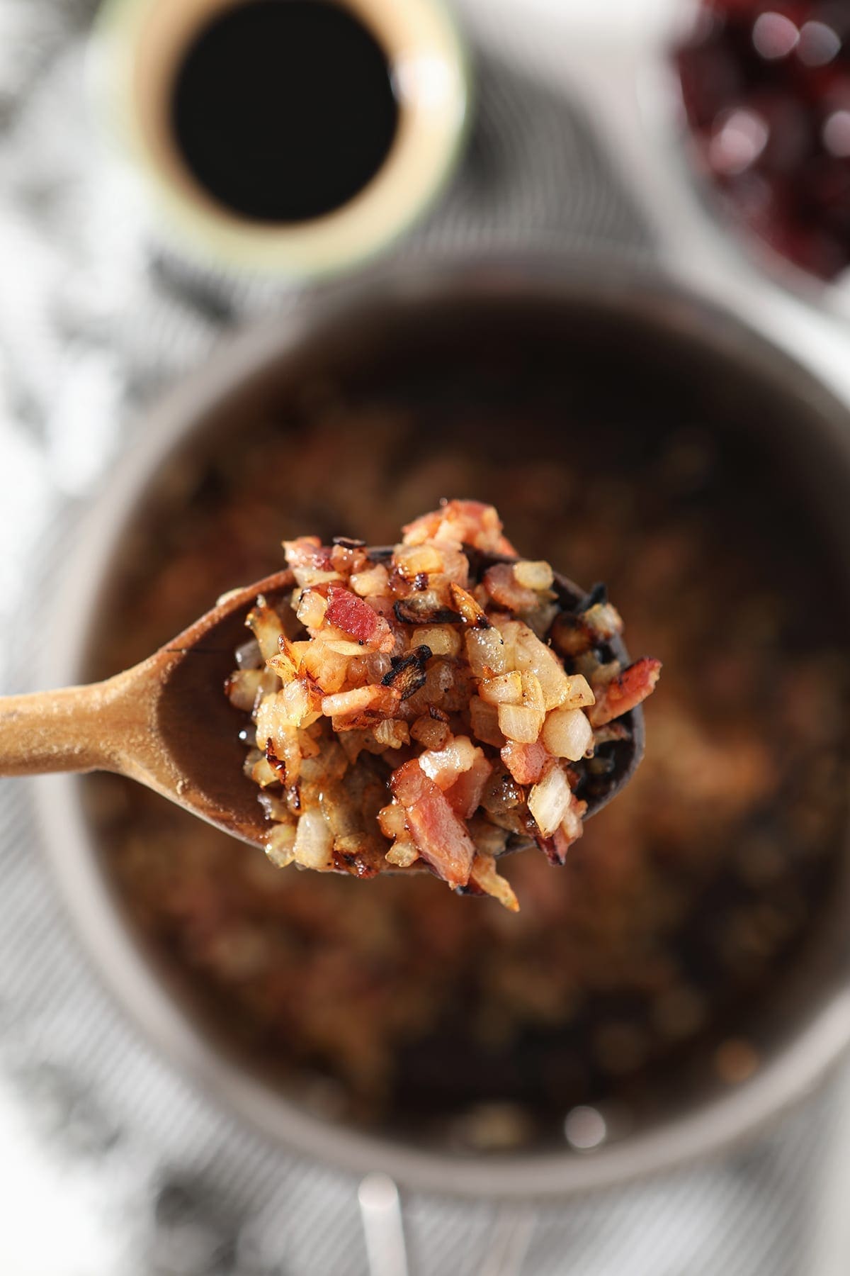 A wooden spoon holds cooked onion and bacon above a saucepan