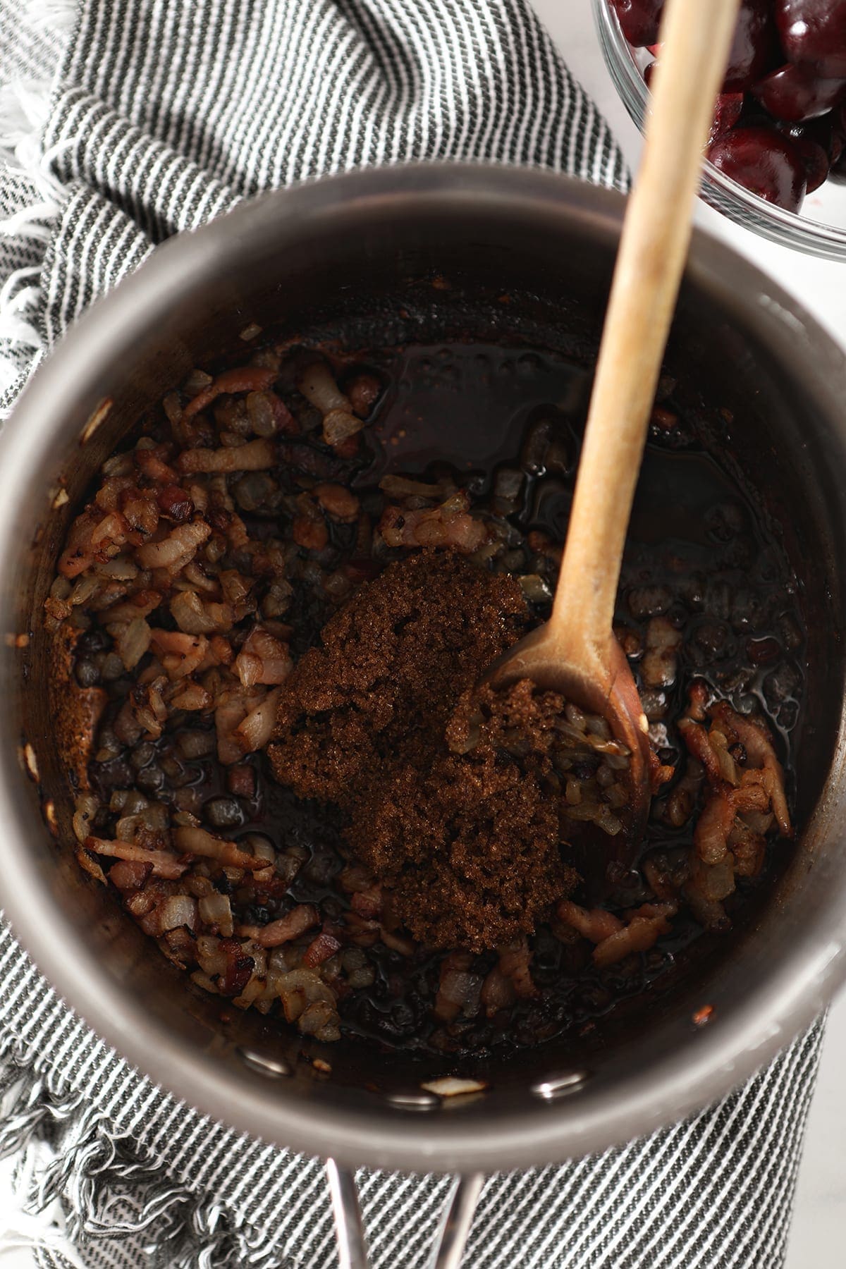 Ingredients for the Bacon Cherry Compote inside a silver saucepan with a wooden spoon before stirring brown sugar in