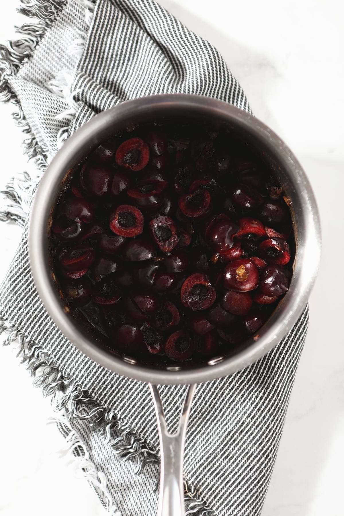 Overhead of a saucepan with fresh halved cherries and other ingredients in it on a grey striped towel