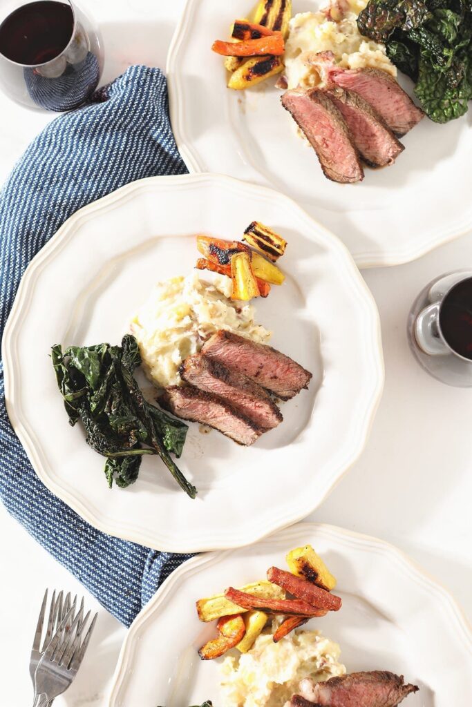 Three plates holding seared steak with mashed potatoes, roasted carrots and greens from above