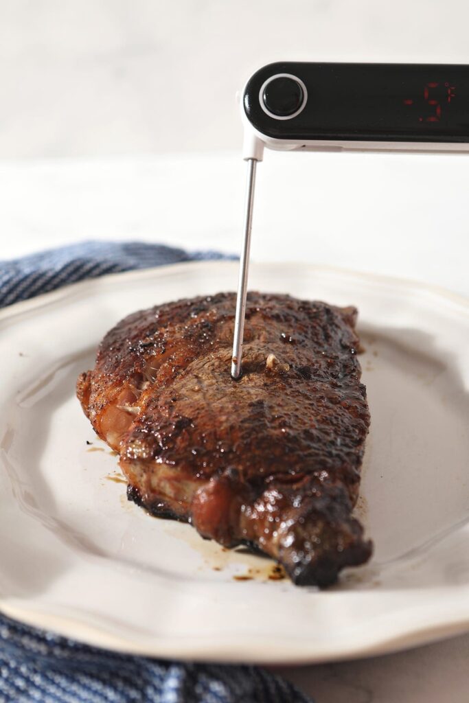 An instant read meat thermometer reads the temp on a ribeye on a white plate