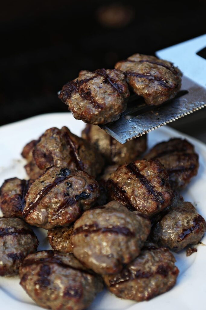 A spatula transfers beef slider hamburgers onto a white plate from the grill