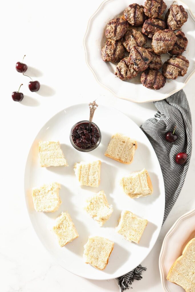 Toasted buns sit with a jar of compote on a white plate with burgers to the side