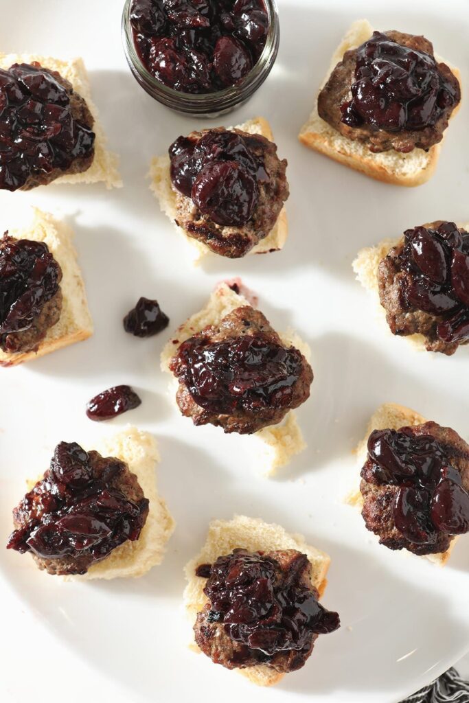 Beef sliders topped with cherry compote sit on a white platter before the tops of the buns are added