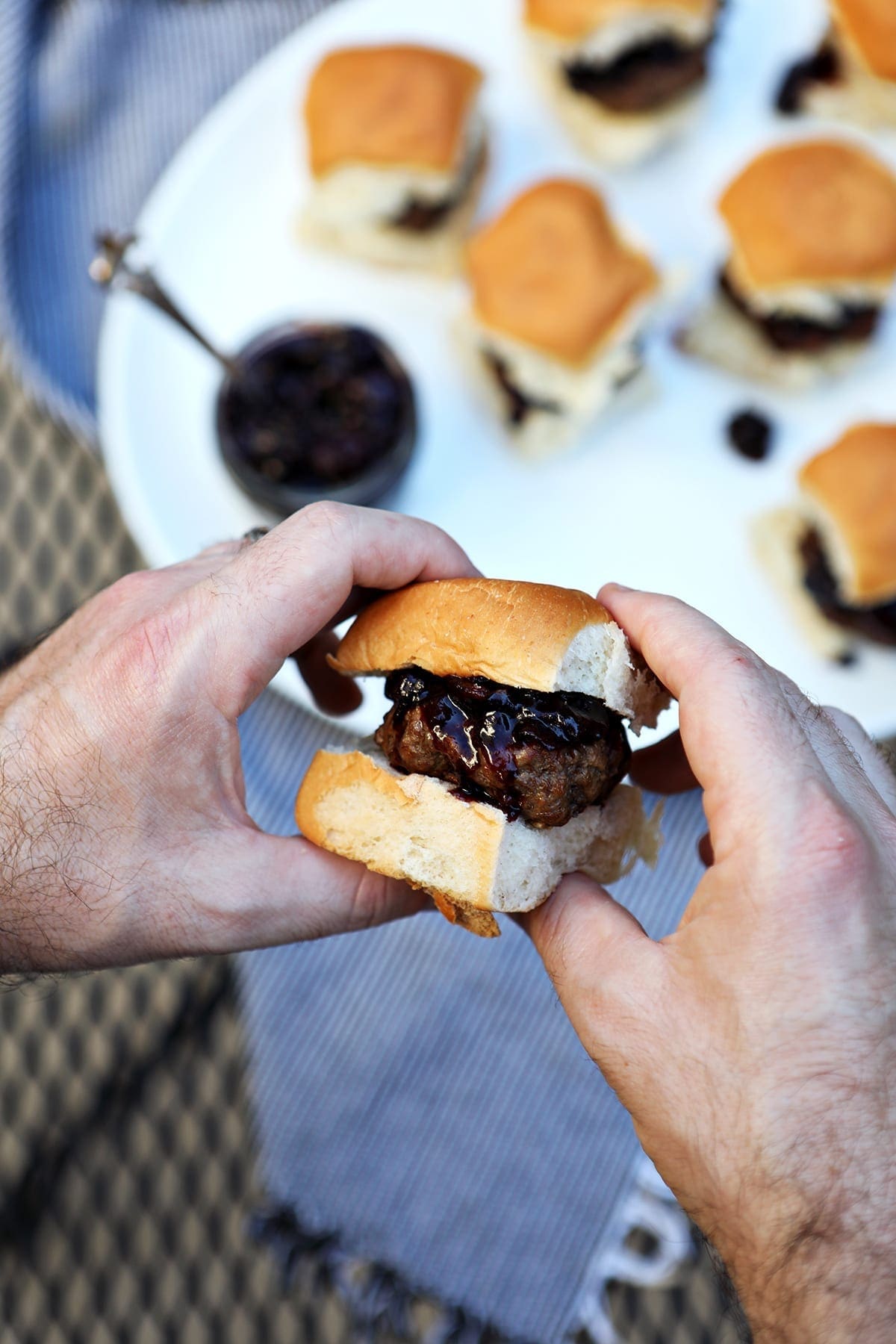 A man holds a slider between his hands with a white platter of other sliders in the background