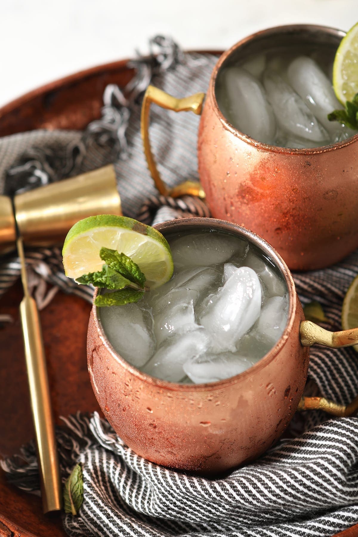 Two full copper mugs garnished with lime and mint sit wrapped in a blue striped towel