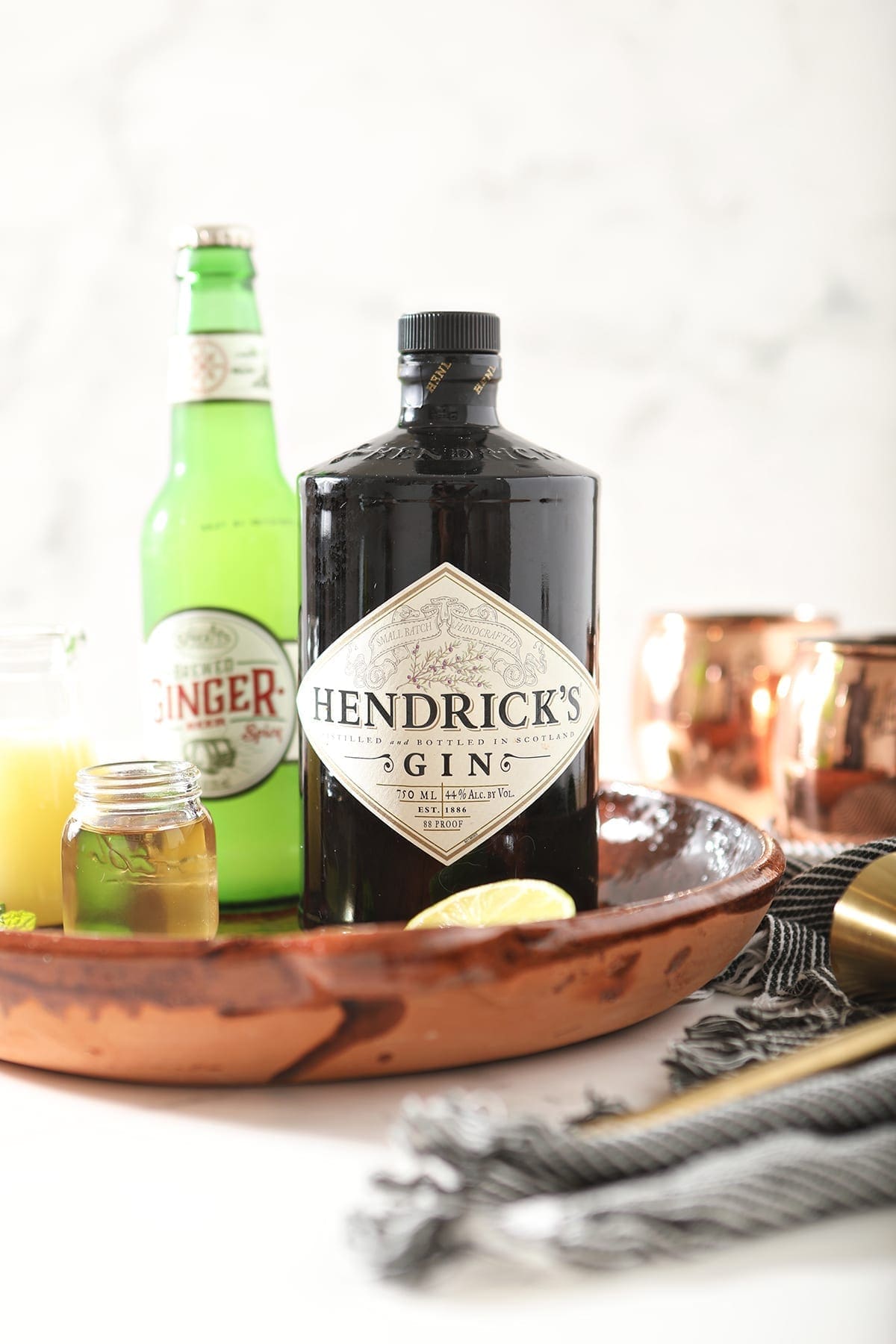 A bottle of Hendrick's gin sits with a bottle of ginger beer, lime juice and simple syrup in a brown serving platter