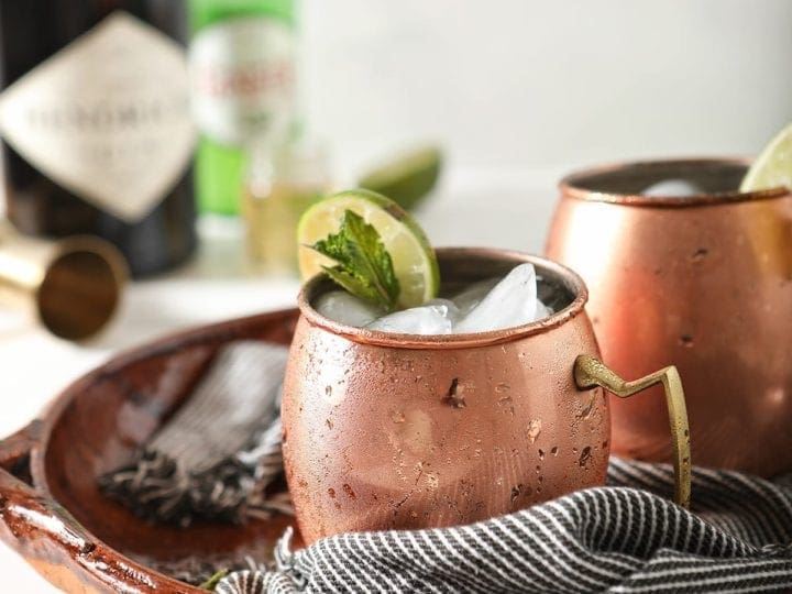 Two copper mugs garnished with lime rounds and mint are shown in a brown serving tray holding Gin Gin Mules