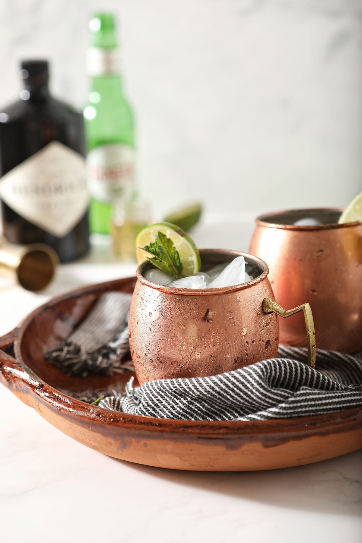 Two copper mugs garnished with lime rounds and mint are shown in a brown serving tray holding Gin Gin Mules