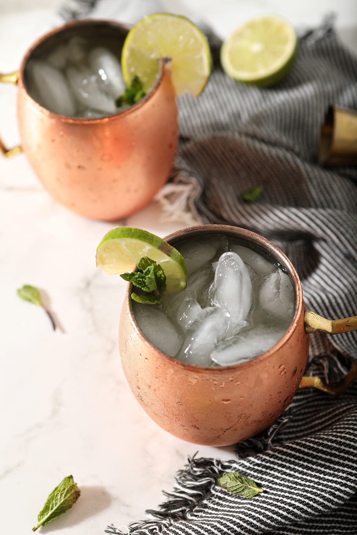 Two Gin Gin Mules in copper mugs on a marble surface sit next to a blue striped towel, lime rounds and mint leaves