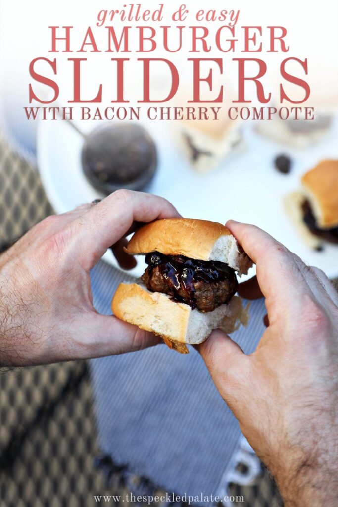 A man holds a slider between his hands with a white platter of other sliders in the background with text "grilled & easy hamburger sliders with bacon cherry compote"