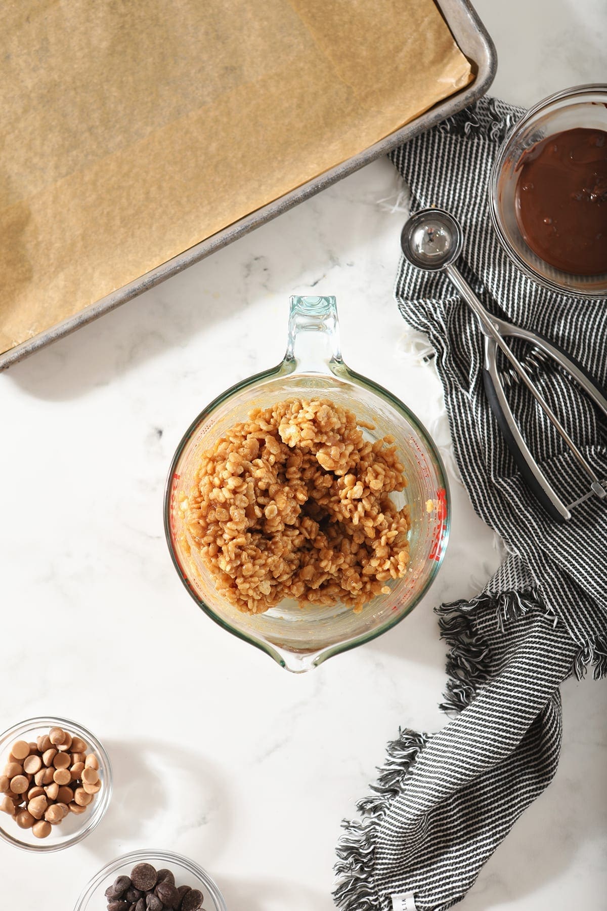 A liquid measuring cup holds the no bake cookie dough while surrounded by a scooper, butterscotch chips and the chocolate drizzle on marble