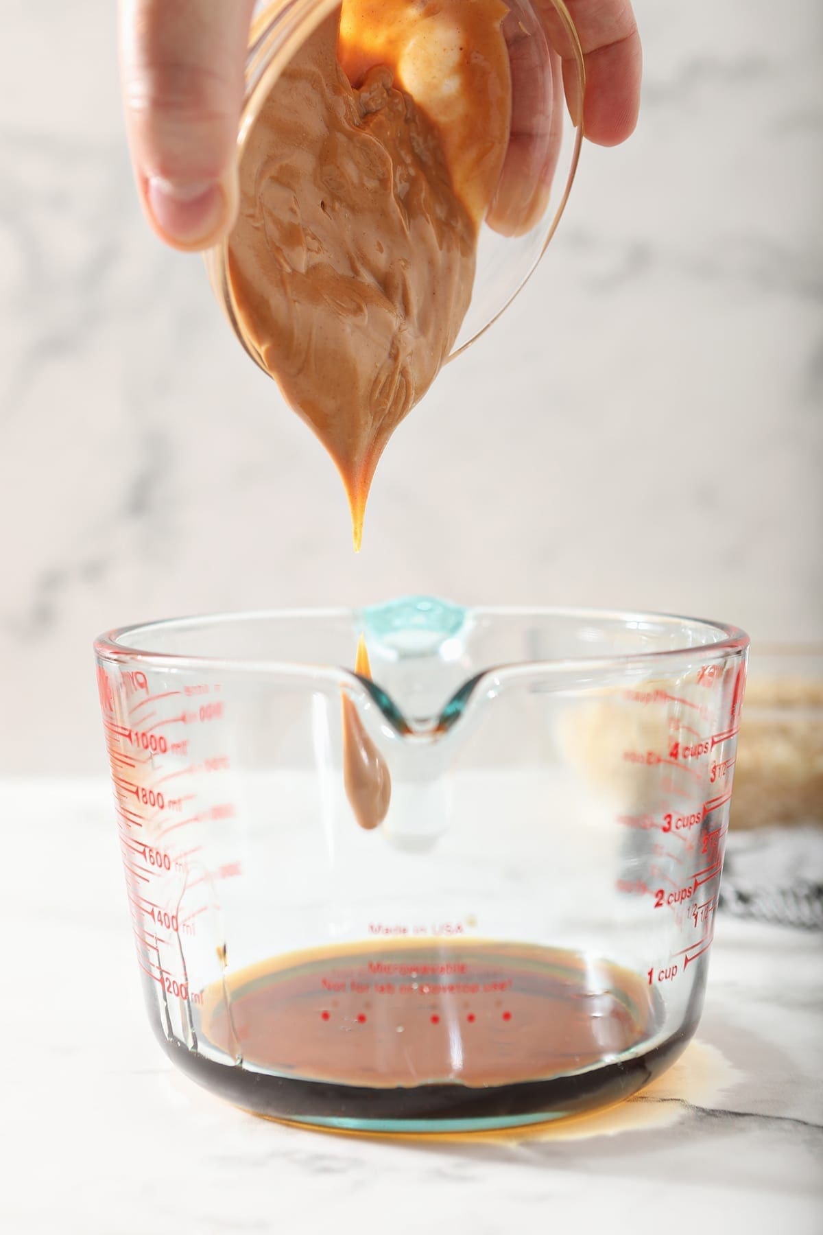 Peanut butter is dolloped into a clear glass measuring cup with brown liquid already in it