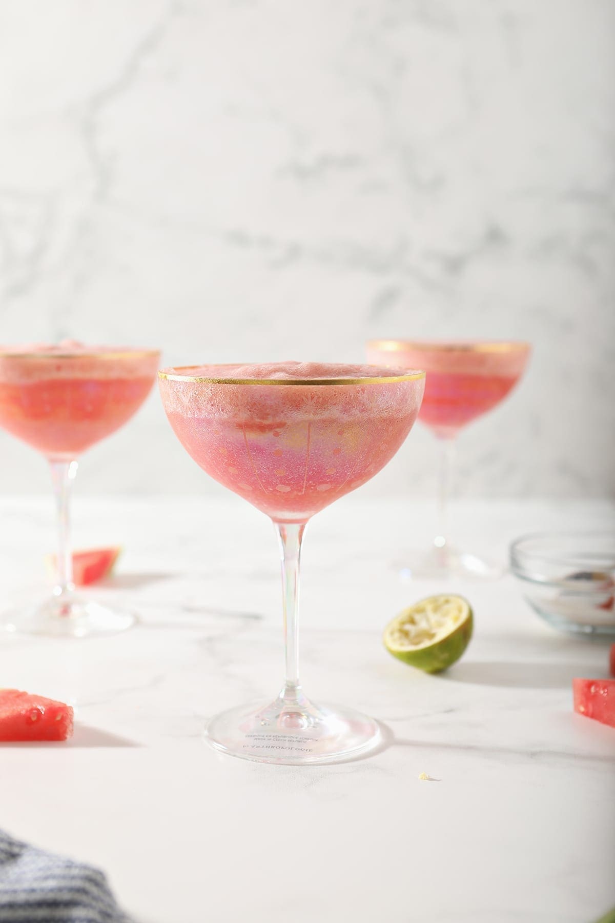 Three coup glasses hold a pink slush drink, surrounded by watermelons and limes on a marble surface