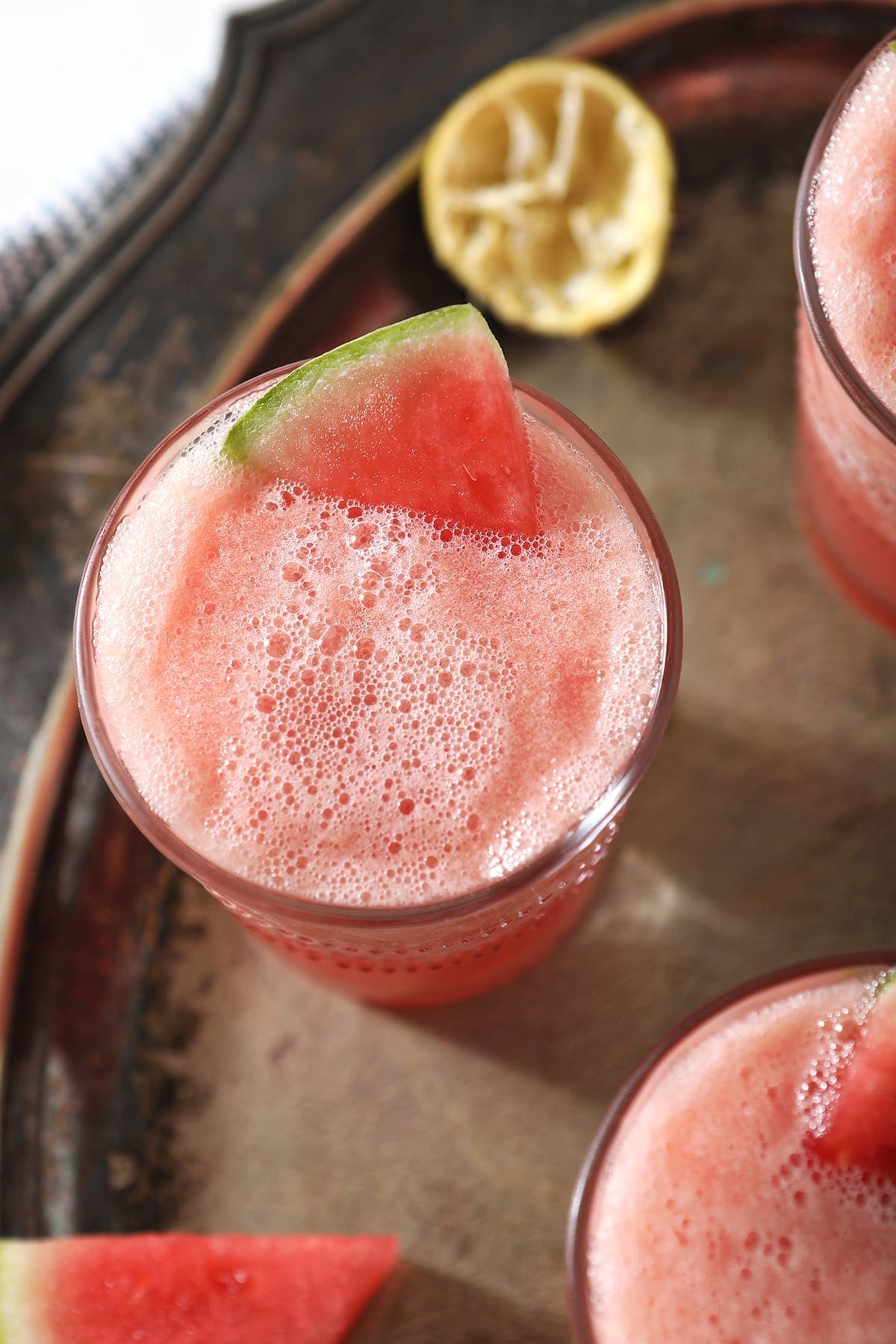 Overhead of a slushy pink drink inside three glasses, garnished with watermelon, on a silver platter