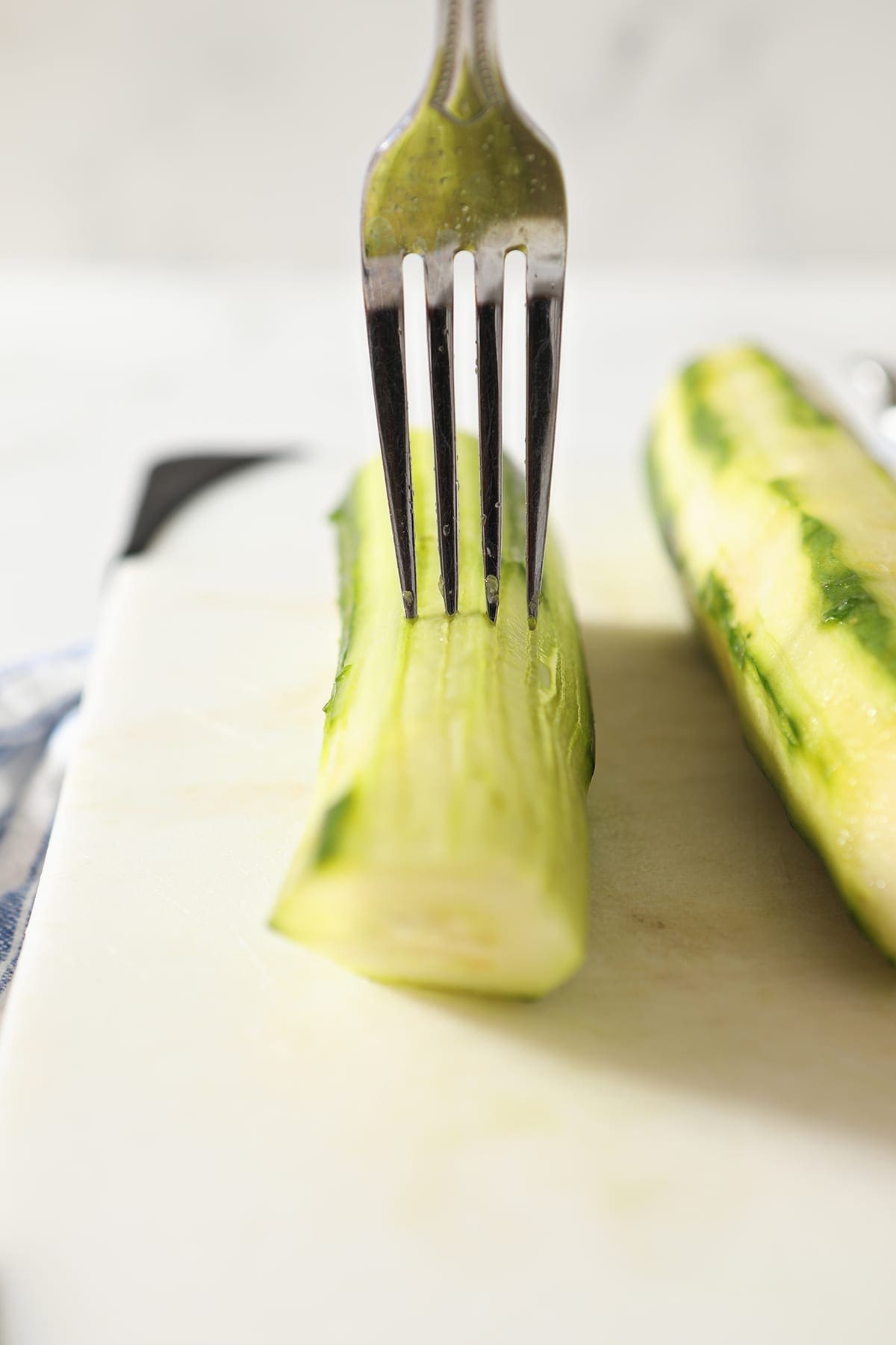 A fork makes indentions onto the top of a peeled cucumber on a white cutting board