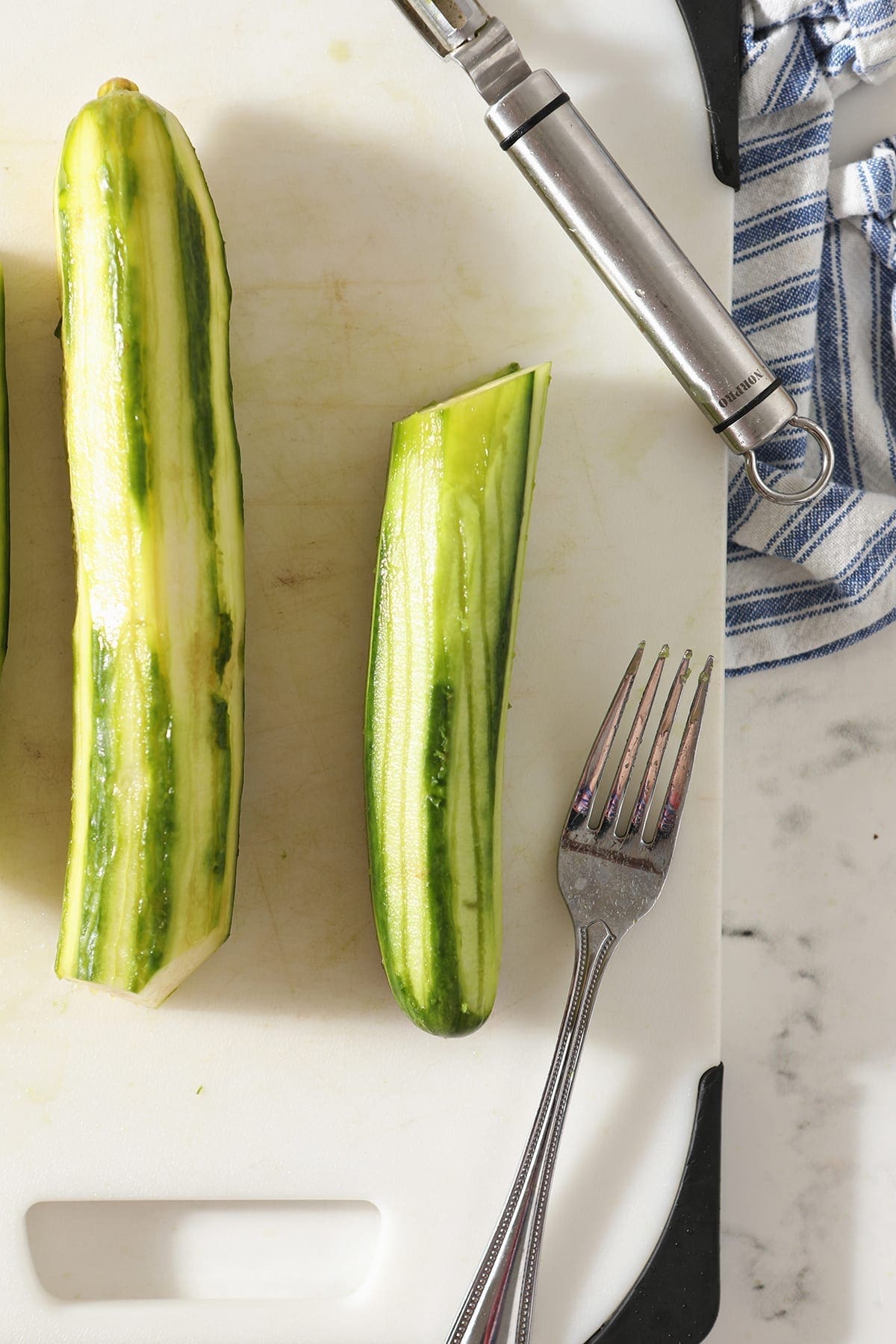 Two cucumbers, peeled and marked with a fork, sit on a white cutting board with a silver fork and a peeler