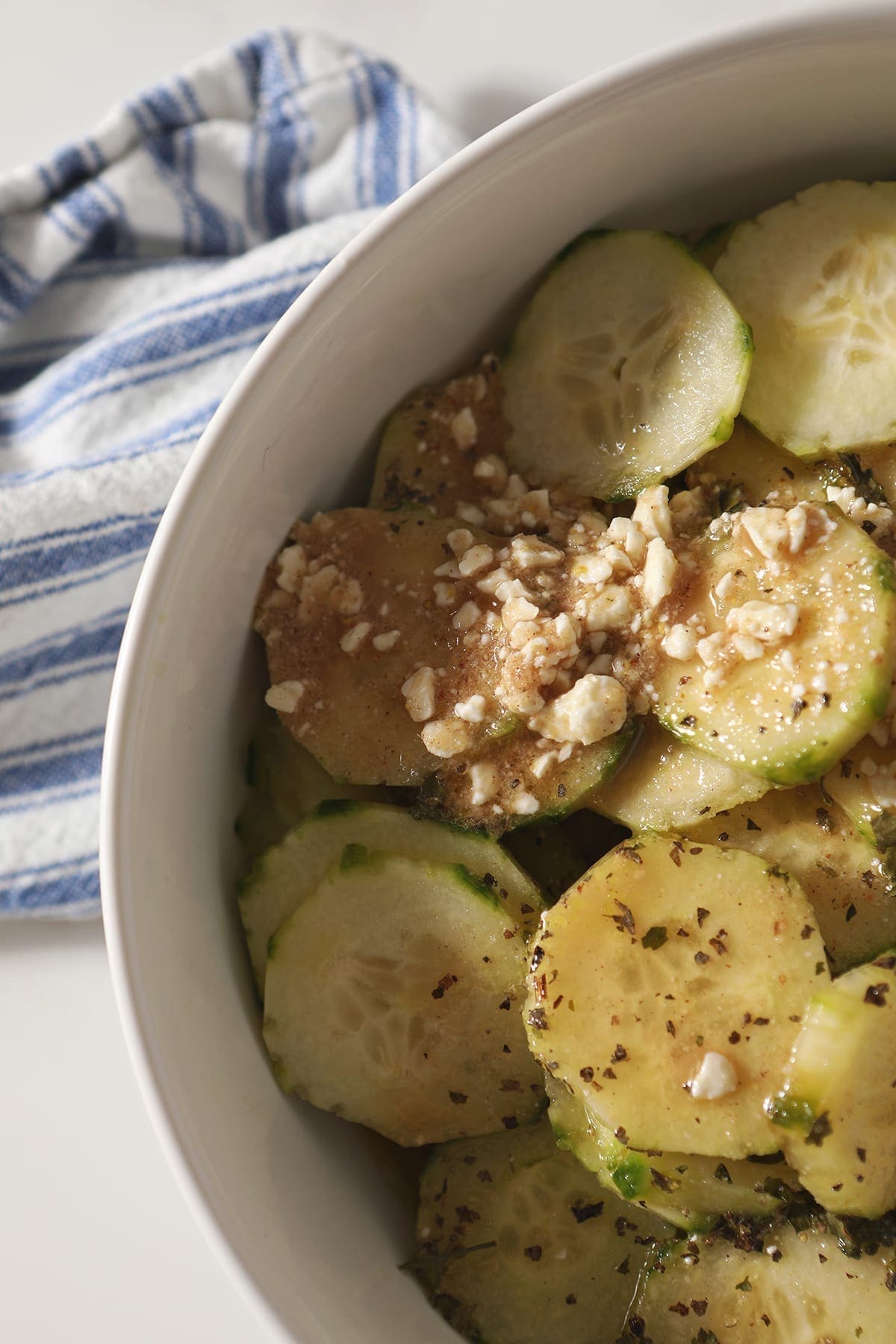 Close up of cucumbers and salad dressing in a white bowl on top of a blue striped towel