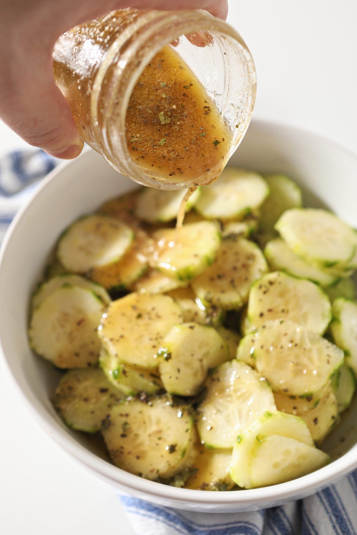 A vinaigrette pours on top of a cucumber salad in a white bowl