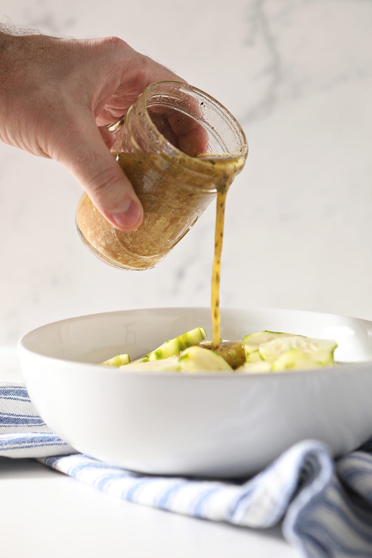 Feta vinaigrette pours on top of cucumber rounds in a white bowl sitting on a blue and white striped towel