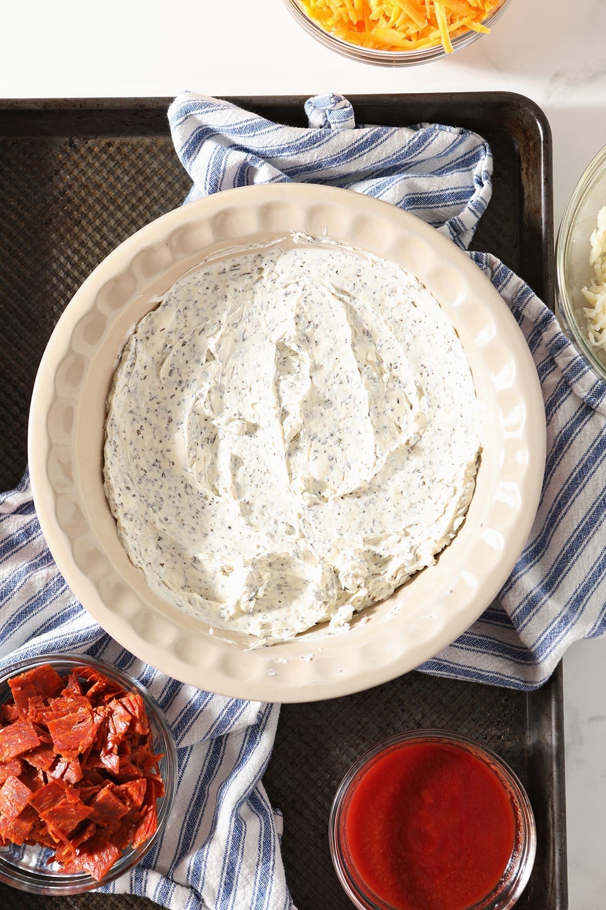 A creamy layer with herbs inside it spread in the bottom of a pie pan, sitting on a blue and white striped towel with other ingredients